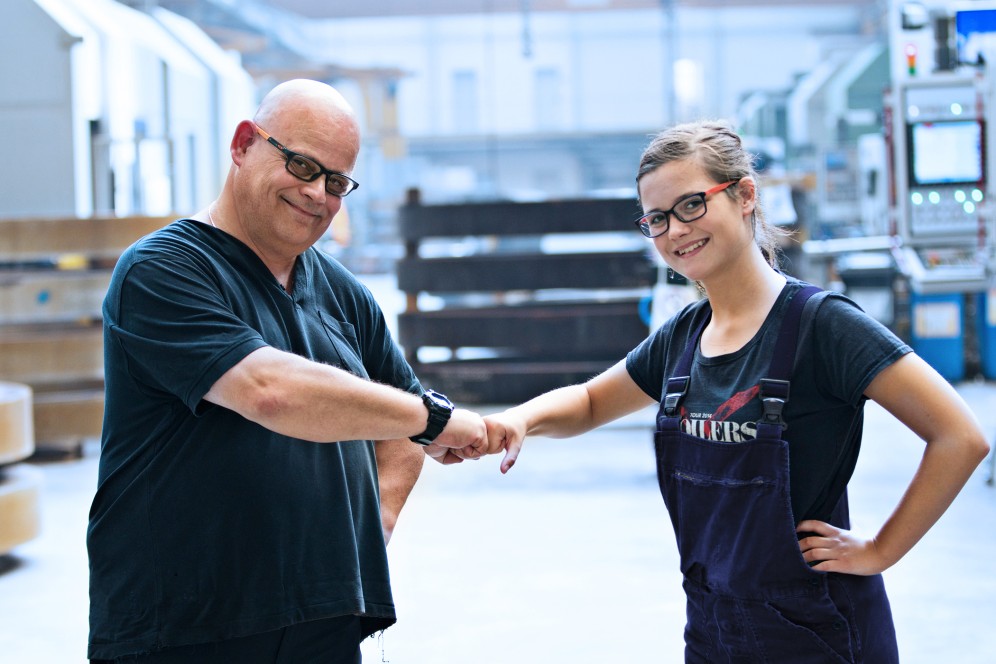 Man with black t-shirt, glasses and bald head and a girl with glasses and blond hair, both look smiling at the camera and give each other fists.