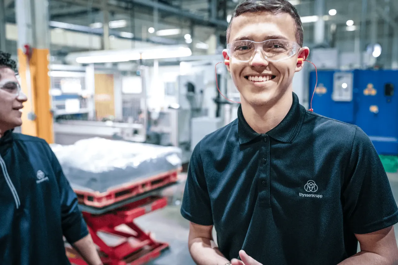 Junger Industrie-Auszubildender mit Schutzbrille und schwarzem Poloshirt steht lächelnd in einer modernen Produktionshalle vor Maschinen und Fertigungsanlagen.