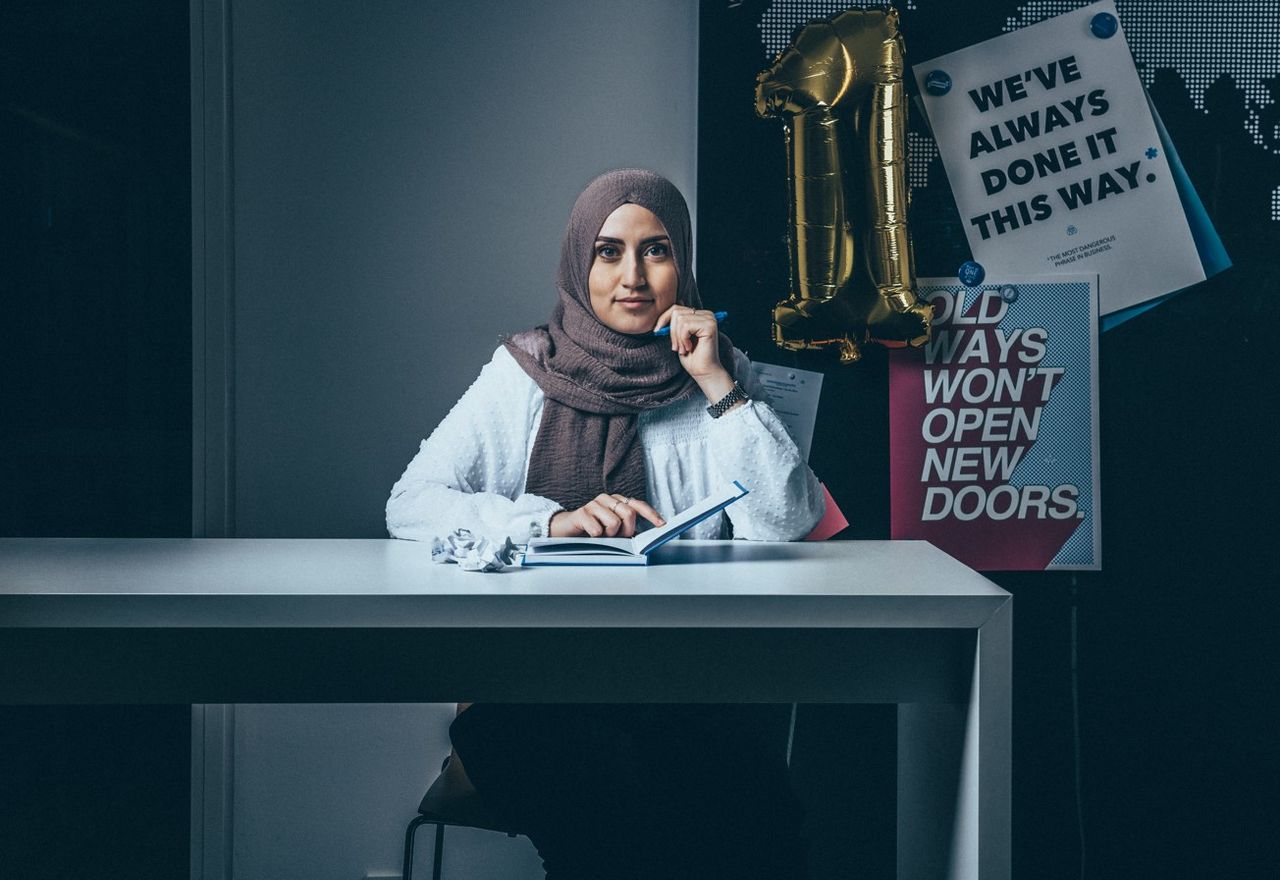Junge Frau sitzt an einem Tisch mit geöffnetem Notizbuch, blickt selbstbewusst in die Kamera. Im Hintergrund inspirierende Poster und eine goldene Ballonzahl Eins.