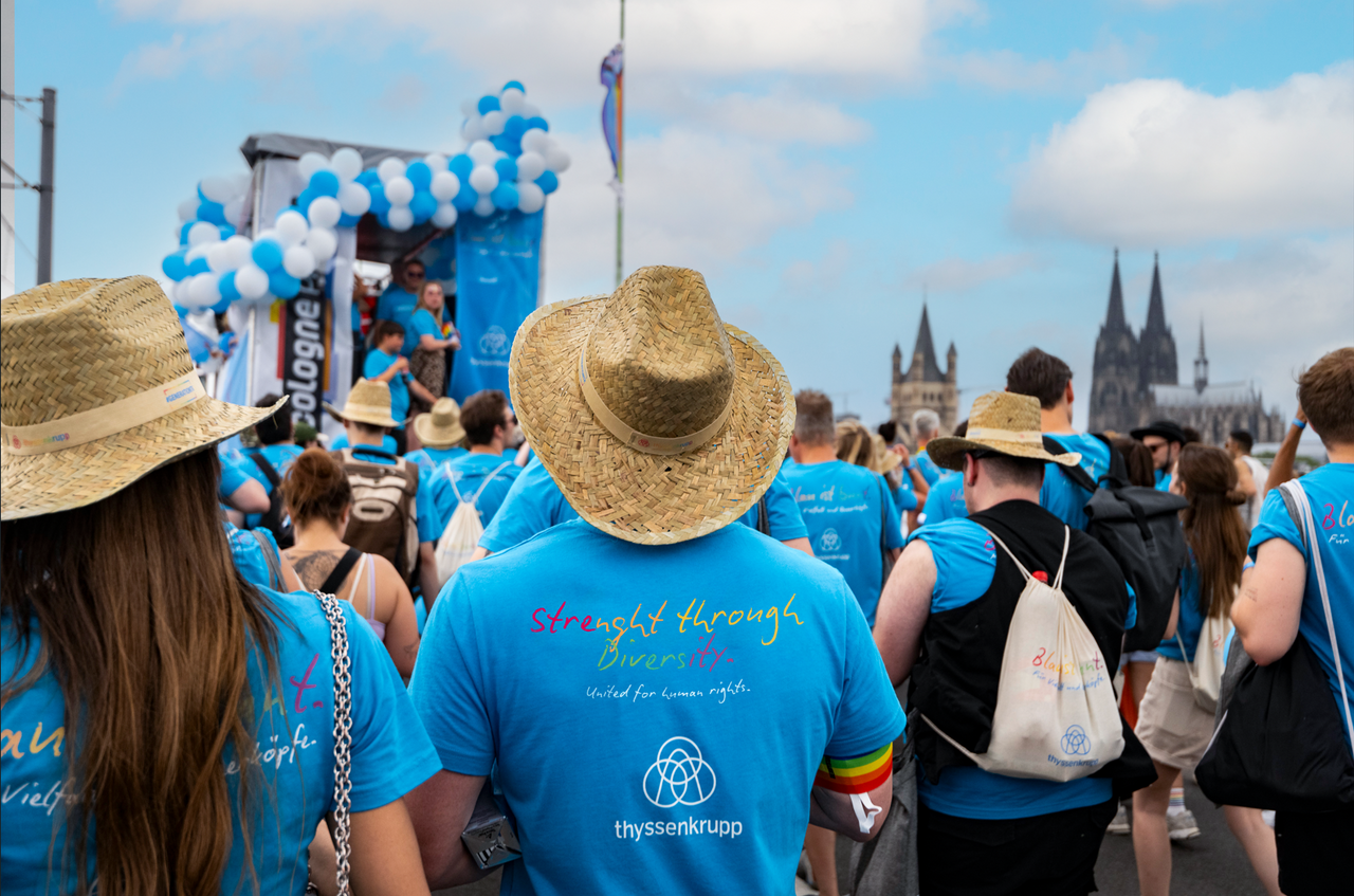 A crowd of people in matching blue shirts and straw hats participating in a public event (CSD), with a stage decorated with balloons and the Cologne Cathedral in the background.
