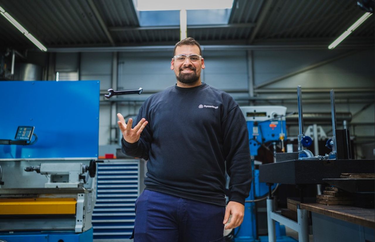 Confident young man in a workshop standing in front of industrial machines, wearing safety glasses and a dark work shirt.