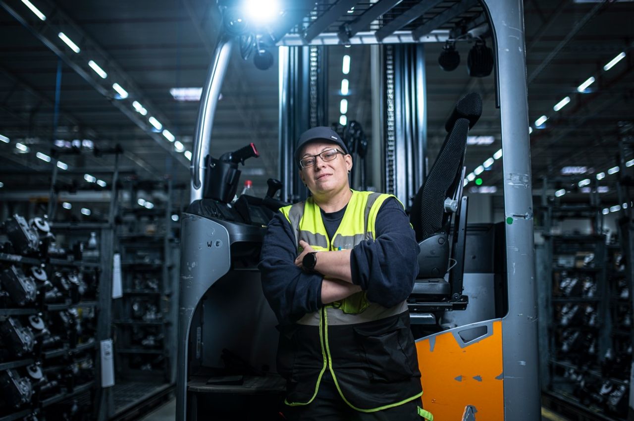 Gabelstaplerfahrerin mit Warnweste und Basecap steht selbstbewusst mit verschränkten Armen vor seinem Fahrzeug in einem großen Lager.