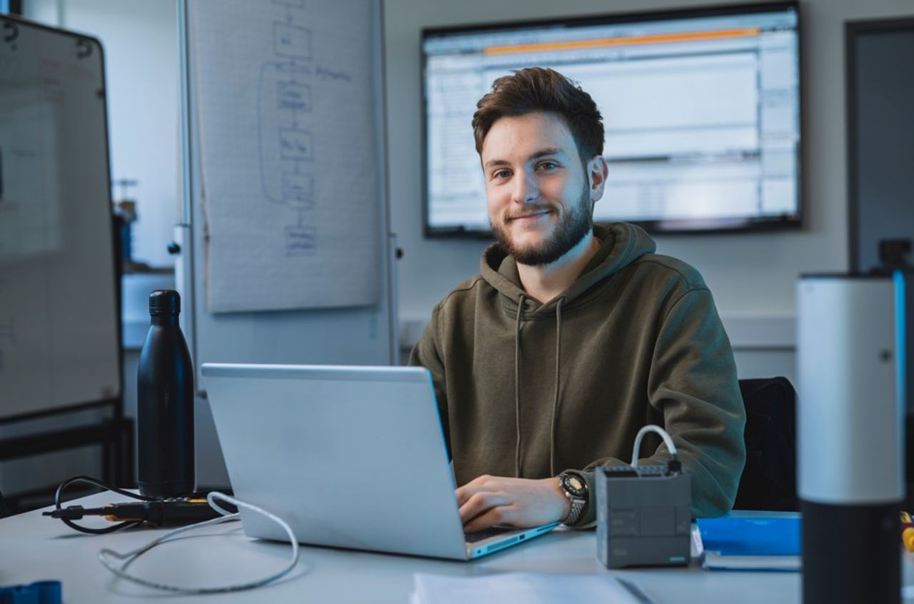 A young man smiling while sitting at a desk with a laptop in a modern office; in the background are technical diagrams and screens.