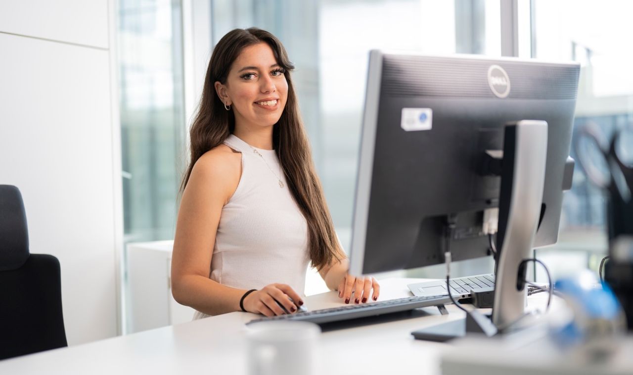 Young woman smiling while sitting at a desk in a bright modern office, working on a desktop computer.